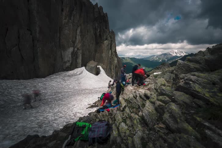 A timelapse of a group of hikers and mountaineers setting up camp on a rocky and snowy mountain pass under a dramatic stormy sky