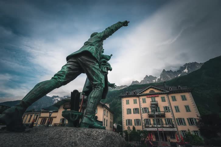 A low-angle timelapse shows the historic Balmat and Saussure statue pointing toward the mountains as clouds move over the busy town square of Chamonix, France