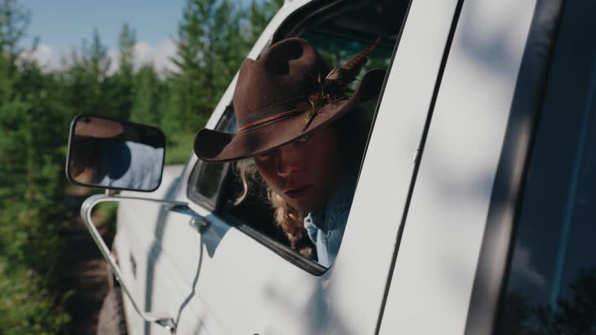 A serious young woman in a cowboy hat stares intently at the camera while sitting in her vintage white pickup truck on a sunny day in the woods