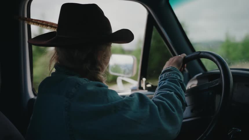 A moody shot of a woman in a wide-brimmed hat driving her pickup truck through a rural, wooded landscape