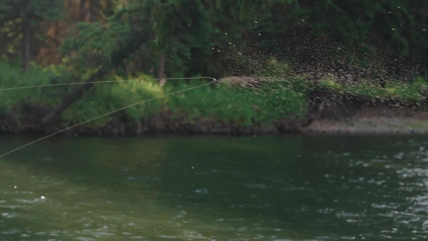 A fly fishing line whips through the air in extreme slow motion, spraying droplets of water over a calm river