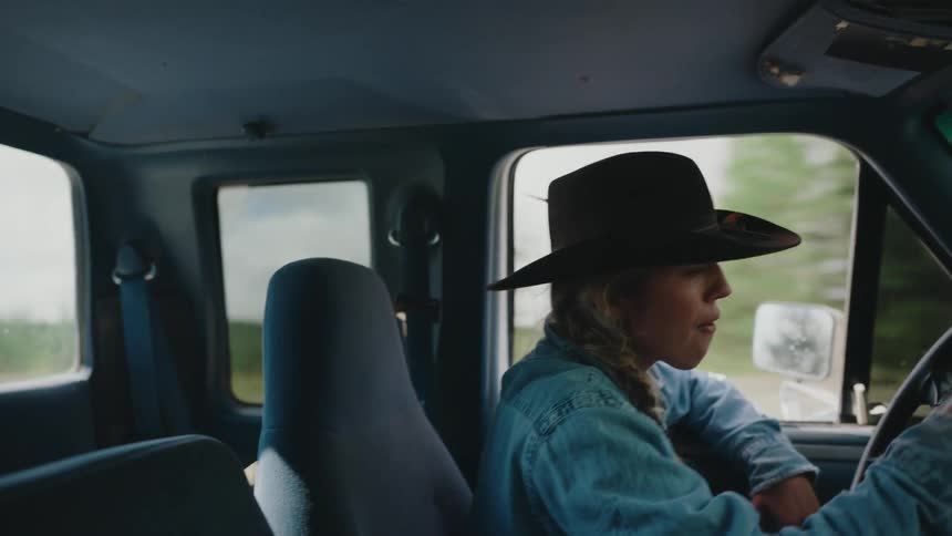A young woman wearing a cowboy hat drives her pickup truck down a rural road, seen from inside the cab