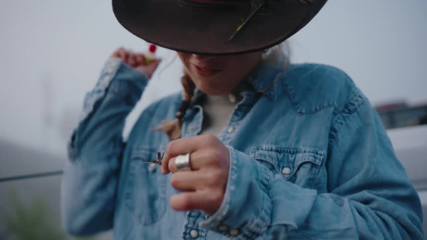 Close up on a woman in a wide-brimmed hat carefully preparing a colorful fishing lure on a foggy day