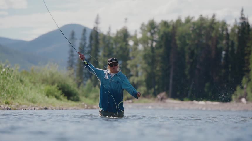 A woman fly-fishes in slow motion while standing in a scenic mountain river on a sunny day
