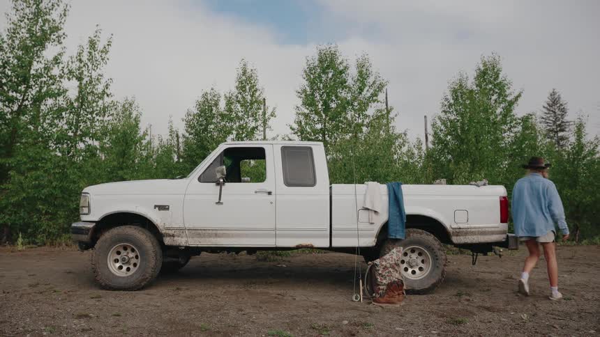 A young woman in a hat walks alongside her white pickup truck on a dirt road, preparing for a fishing trip in the woods