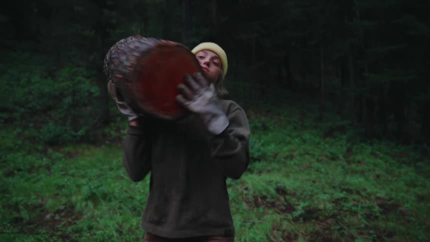 A strong woman in a yellow beanie lifts a heavy log from the forest floor and carries it on her shoulder in slow motion