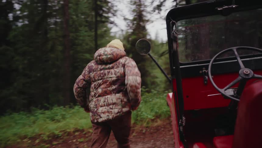 An adventurer in a camo puffer jacket gets out of a vintage red off-road vehicle to explore the deep forest in slow motion