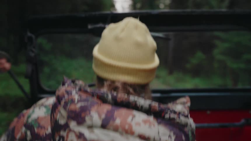 Back shot of a traveler in a yellow beanie riding in a bumpy off-road vehicle on a trail through a lush green forest