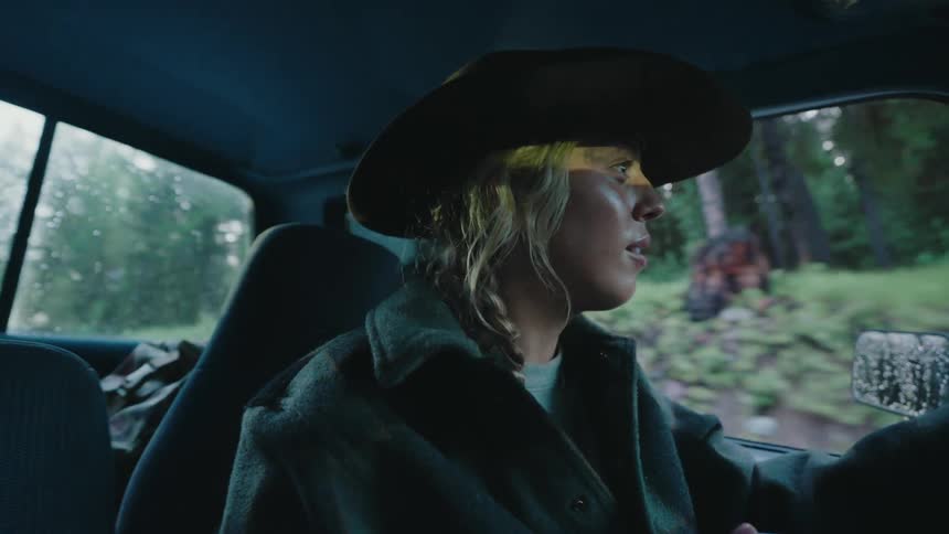 A woman in a cowboy hat drives her truck through a moody, rainy forest with a determined expression