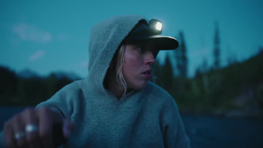 A tense woman wearing a hoodie and a headlamp looks over her shoulder while paddling a boat on the water at dusk
