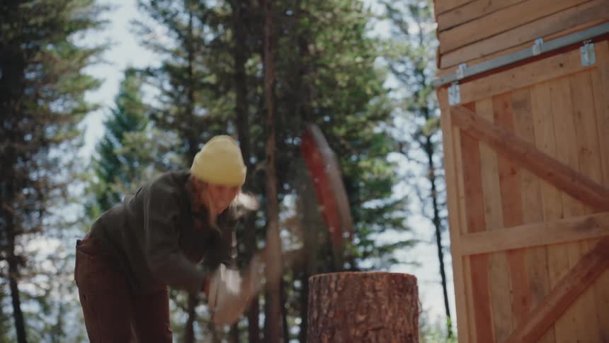 A self-sufficient woman in a beanie and work gloves powerfully splits a large log with an axe in a forest setting