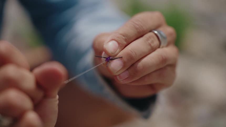 Close-up slow motion shot of a person's hands tying a purple fishing fly onto a clear line