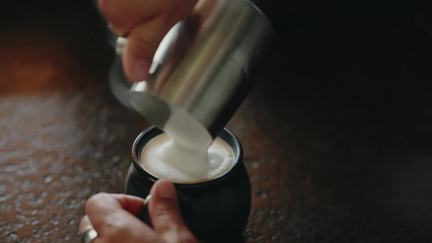A barista slowly pours steamed, frothed milk from a silver pitcher into a dark mug of coffee
