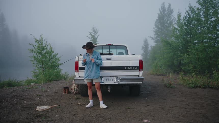 A woman in a cowboy hat prepares her fishing gear while leaning against her white pickup truck on a foggy morning by the river
