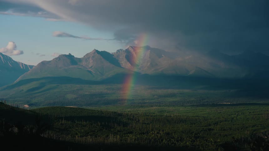 A stunning rainbow emerges from dramatic storm clouds over a vast mountain landscape and lush green forest