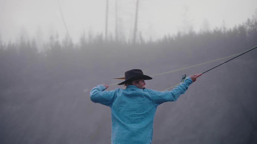 A lone angler in a cowboy hat gracefully casts a fly fishing line into the thick, mysterious morning mist in slow motion