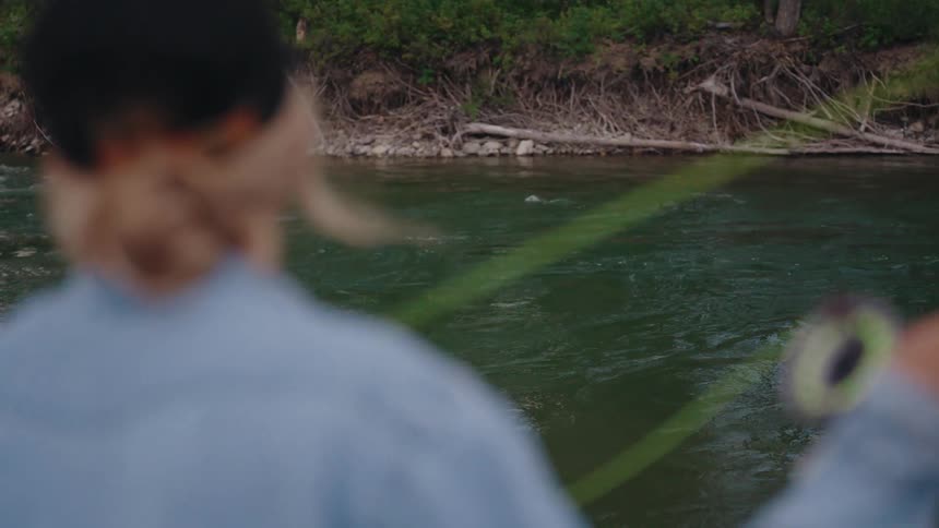From an over-the-shoulder perspective, a fly-fisher gracefully casts their line into the river in super slow motion