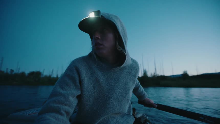 A determined woman wearing a headlamp rows her boat down a river in the wilderness during twilight