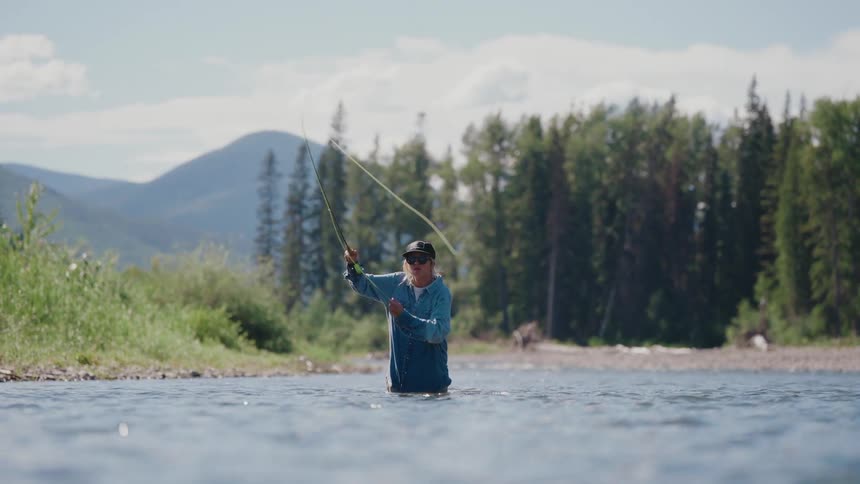 A skilled angler practices fly fishing in the middle of a beautiful river surrounded by mountains and forests in slow motion
