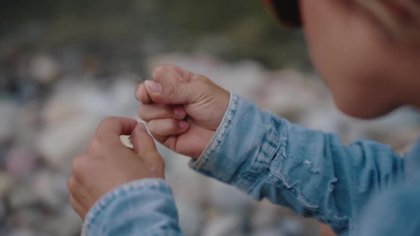 Close up shot of a woman tying a fly lure onto her fishing line with a rocky riverbank in the background
