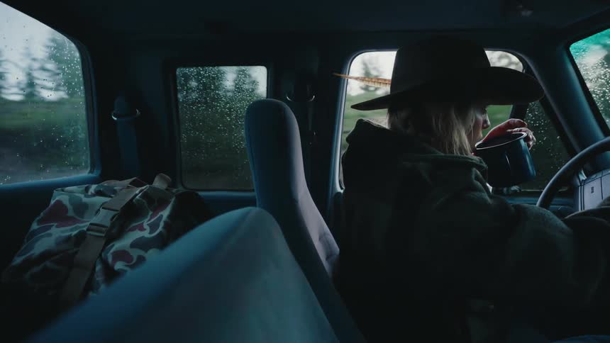 A woman wearing a hat with a feather drives an old truck on a moody, rainy day, taking a sip from her mug