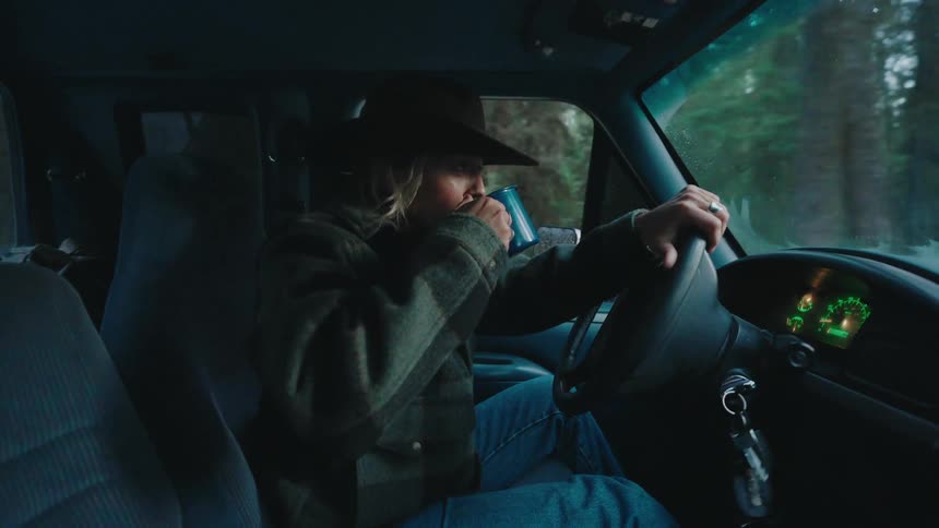 A woman wearing a hat enjoys a warm drink from a mug while driving her pickup truck through a moody forest in slow motion