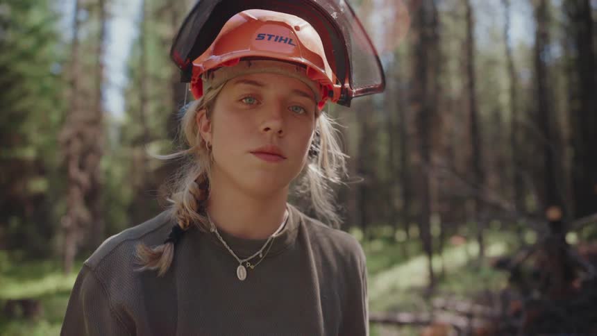 A young female forestry worker wearing a safety helmet slowly looks up with a determined gaze while working in a sunlit forest