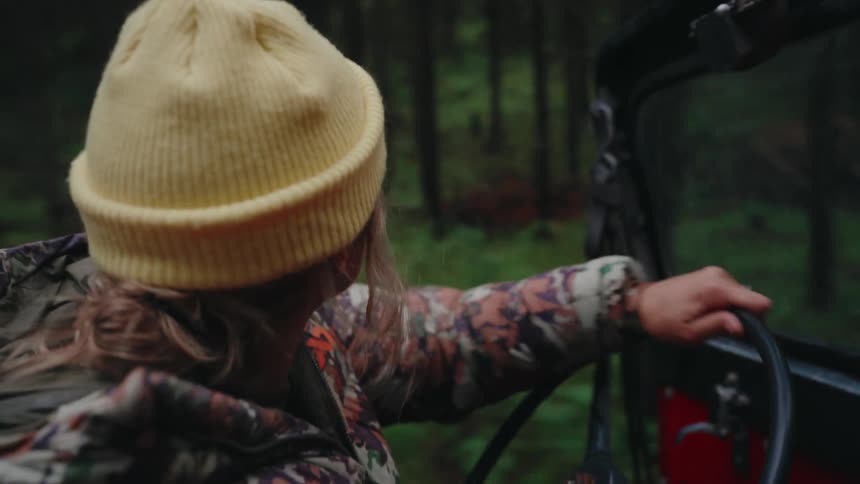 A woman in a yellow beanie and camo jacket drives a rugged vehicle through a bumpy, dense forest