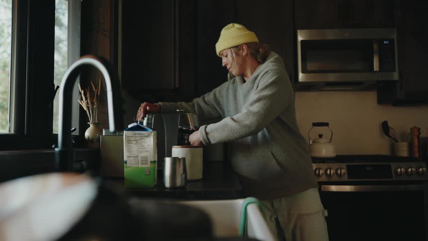 A woman wearing a yellow beanie makes an espresso in a cozy cabin kitchen and pauses to look out the window
