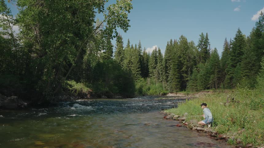 A young boy fishes in a serene mountain river surrounded by a lush green forest on a sunny day