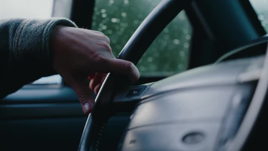 An anxious hand nervously taps on a steering wheel while waiting inside a car on a gloomy, rainy day