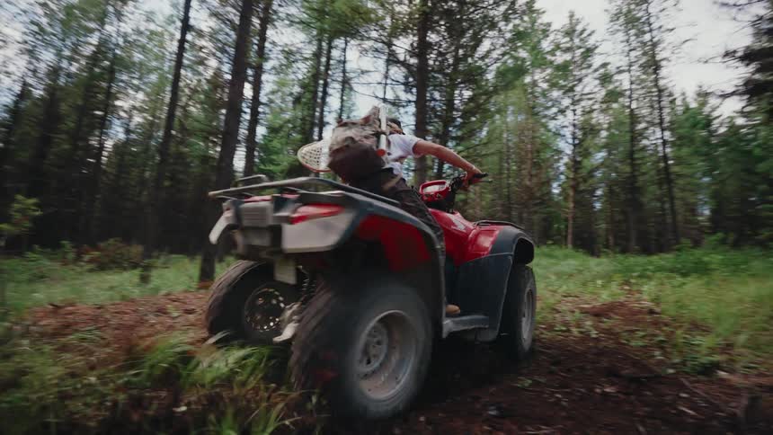 A female hunter with a rifle rides a red ATV away from the camera down a rugged forest trail