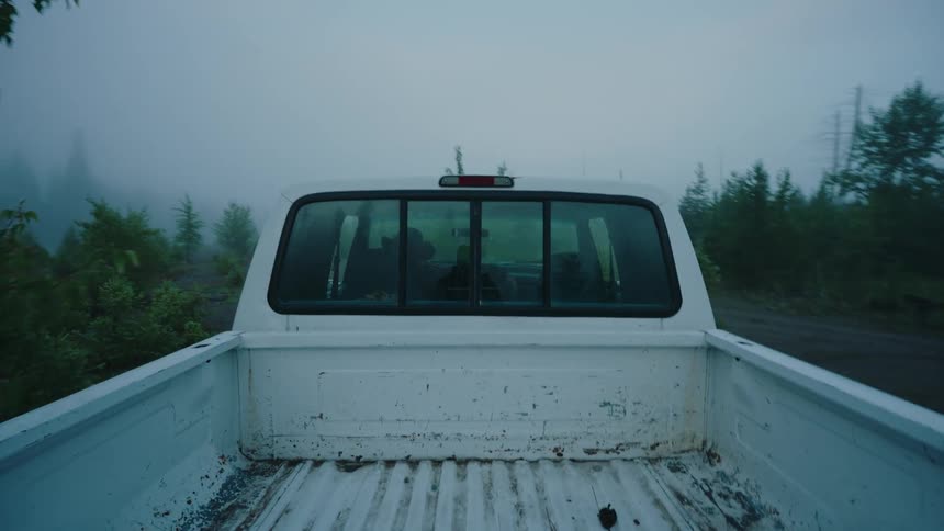 POV shot from the bed of a white pickup truck as it drives backward down a remote and foggy forest road