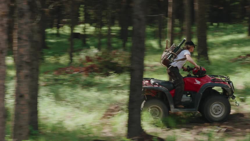 A young adventurer with fishing gear on his back rides a red ATV through a sun-dappled green forest