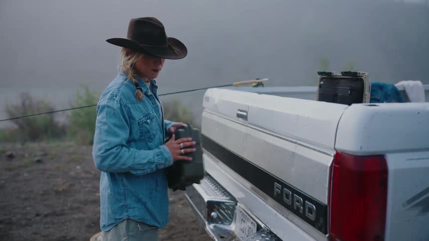 A woman in a cowboy hat selects a fly from her tackle box at the back of her truck and prepares to go fly fishing on a foggy morning