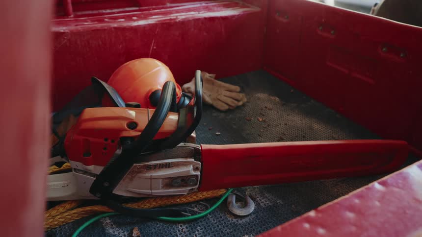 A person loads a chainsaw, hard hat, gloves, and gas cans into the back of a red utility vehicle