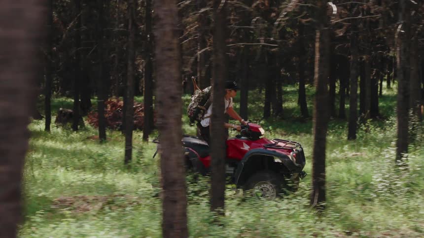 An outdoorsman with a large backpack drives a red ATV through a dense, sunlit forest