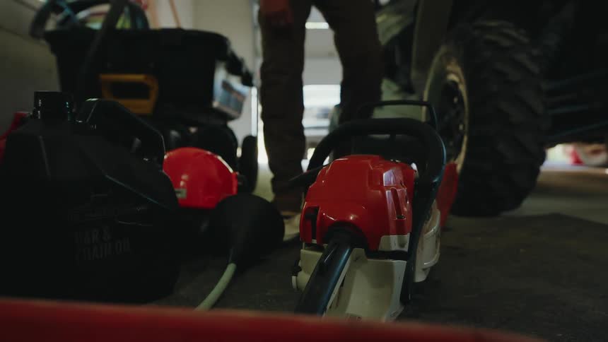A worker walks into a garage to pick up a chainsaw, hard hat, and gas can before heading out for a day of work