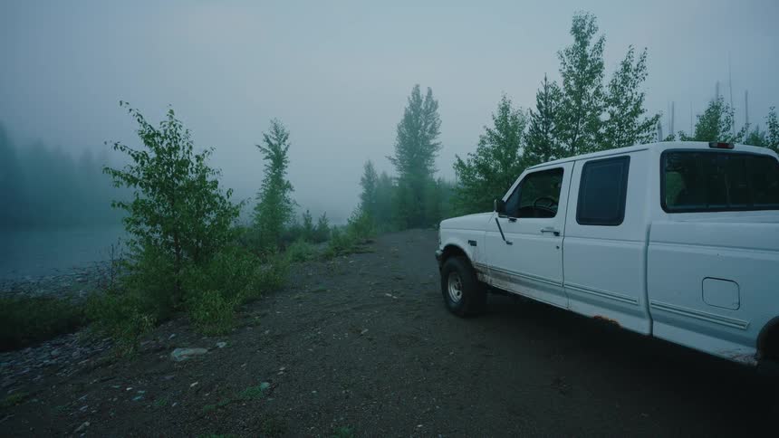 A person wearing a cowboy hat parks their white pickup truck next to a foggy river and gets out