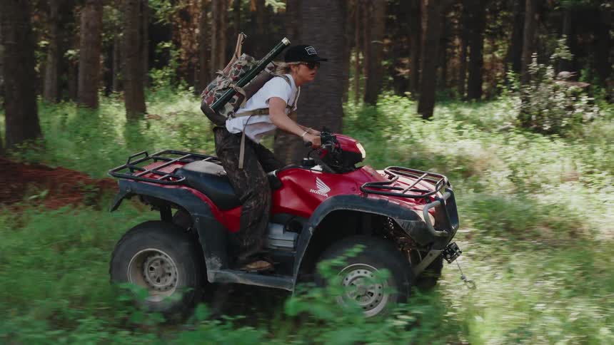 A woman rides a red ATV through a lush green forest with a backpack and fishing gear in slow motion