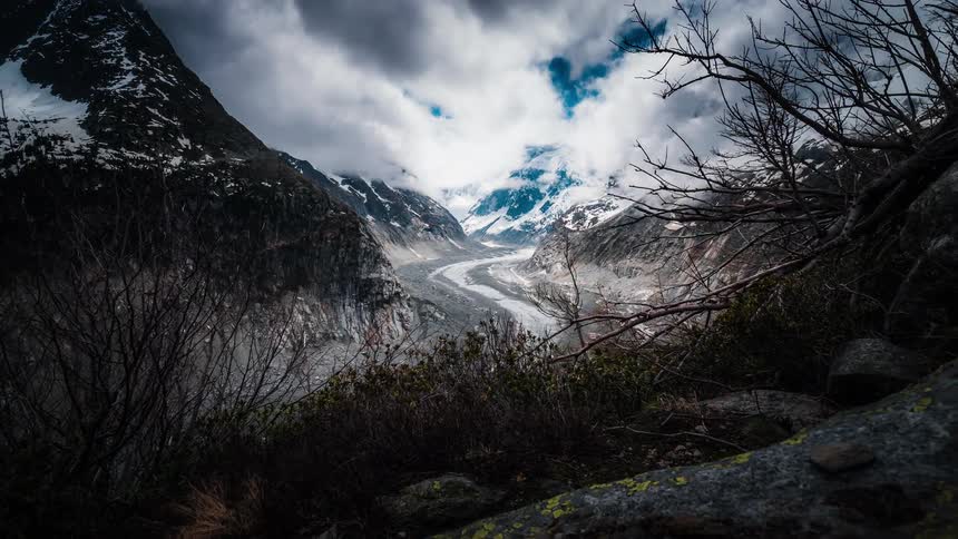 Dramatic timelapse of clouds swirling over a vast glacier winding through a snow-capped mountain valley, revealed from a rocky viewpoint