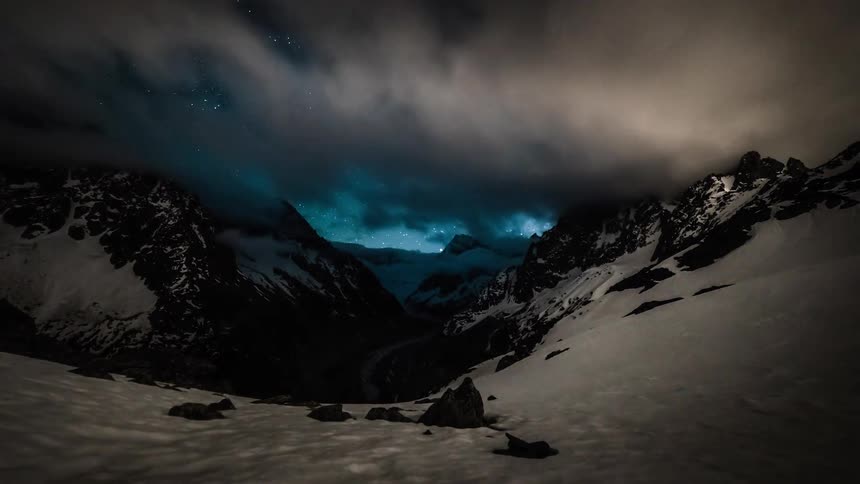 Timelapse of clouds parting to reveal a brilliant, starry night sky and the Milky Way over a snow-capped mountain valley