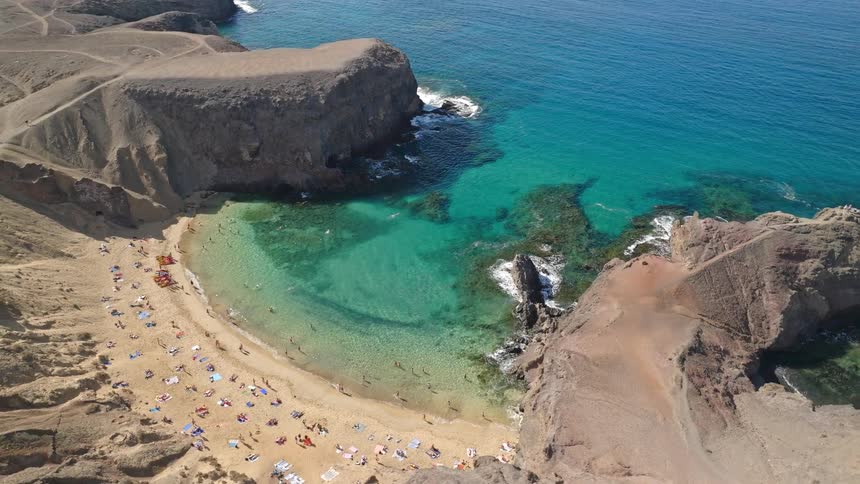 A high-angle aerial view slowly pulls back from a picturesque secluded beach cove filled with sunbathers and swimmers enjoying the turquoise sea