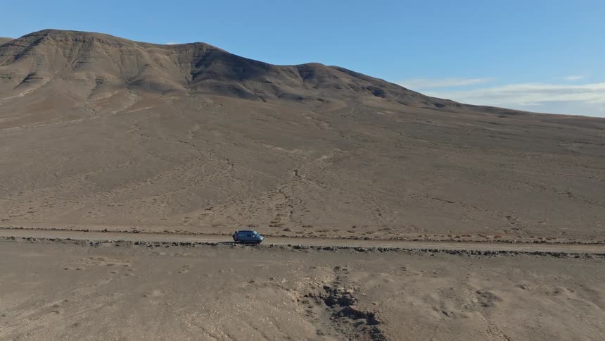 An aerial view shows a lone camper van driving on a remote dirt road through a vast, arid volcanic landscape under a clear blue sky