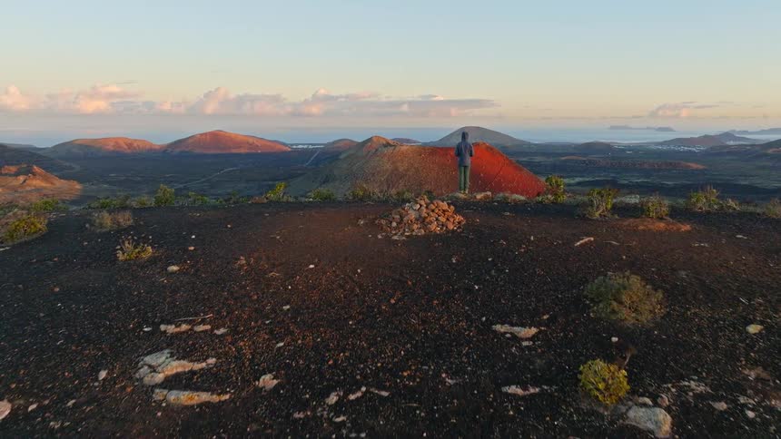 Aerial shot pushes past a lone hiker standing on a desolate hilltop to reveal a breathtaking volcanic landscape at sunrise