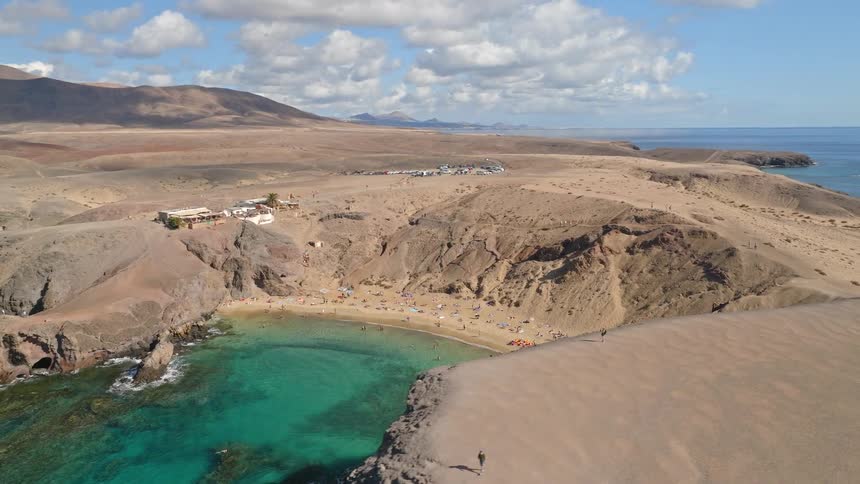 A stunning aerial view pans across a secluded cove with turquoise water and a bustling beach, set against a vast desert landscape