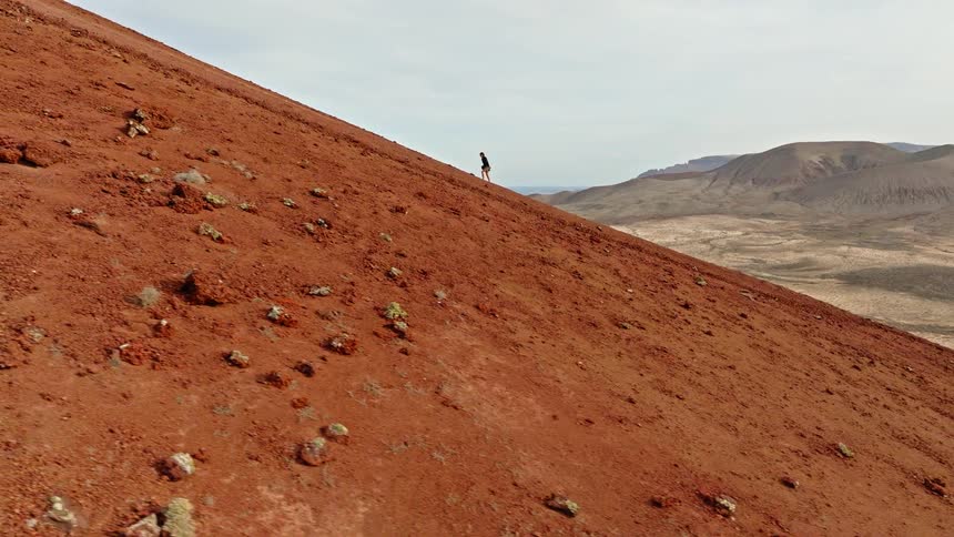 A lone hiker slowly climbs the steep, red volcanic slope of a mountain in a vast desert landscape