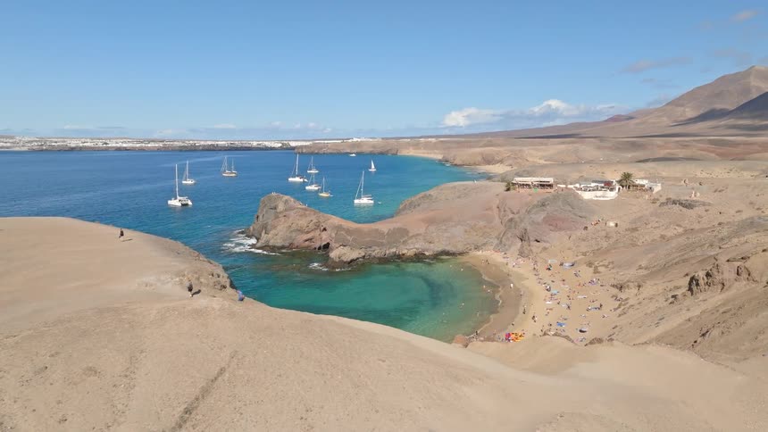 A stunning aerial view reveals a beautiful hidden cove with turquoise water, a sandy beach full of people, and sailboats anchored in the bay