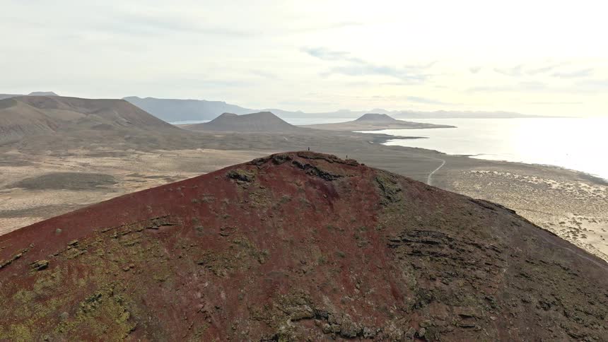 An epic aerial view reveals hikers on the summit of a massive red volcano overlooking the vast coastal landscape