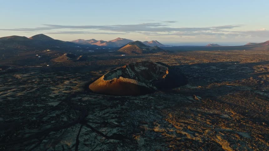 A static aerial shot captures a volcanic crater and vast, rugged lava fields illuminated by the golden light of sunrise